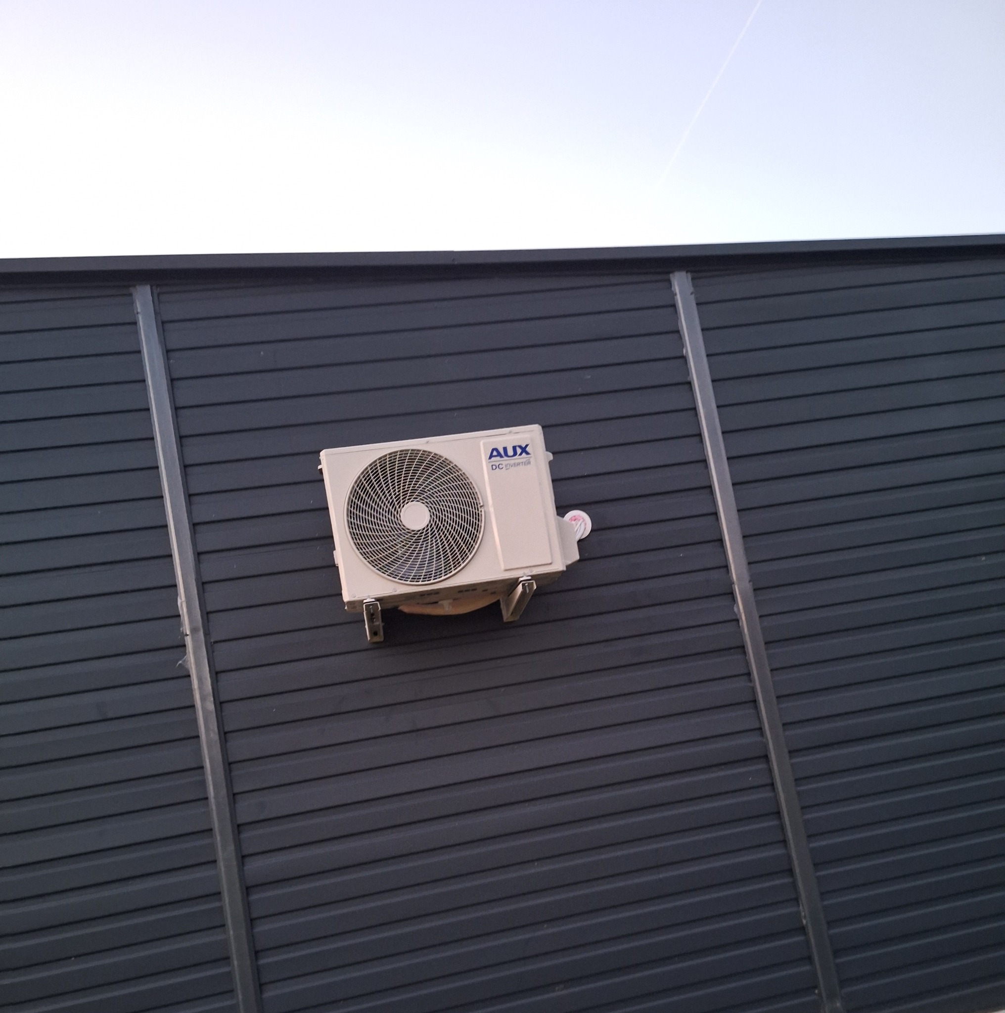 Zamontowana jednostka zewnętrzna klimatyzacji AUX na ciemnej, poziomej elewacji z widocznymi mocowaniami. Ujęcie z dołu, niebo w tle.