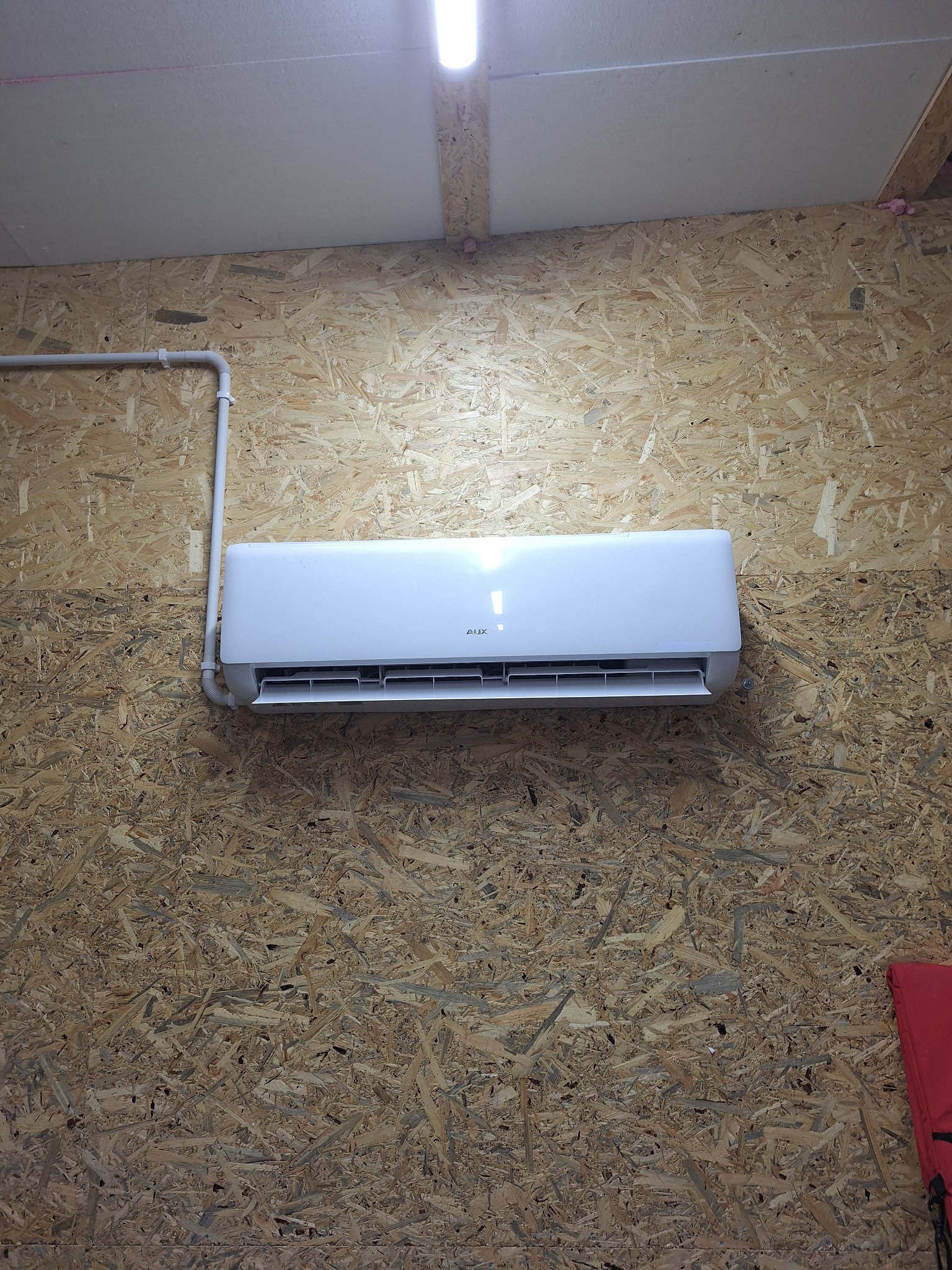 Biały klimatyzator AUX zamontowany na ścianie z płyty OSB, z widoczną instalacją. Oświetlenie sufitowe podkreśla nowoczesny design urządzenia.