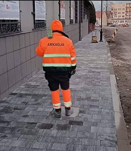 Pracownik w pomarańczowym ubraniu z napisem ARKADJA na plecach, z niwelatorem na ramieniu, stoi na nowo ułożonej szarej kostce brukowej w Sitkówce-Nowinach. Widoczny fragment budynku w tle.