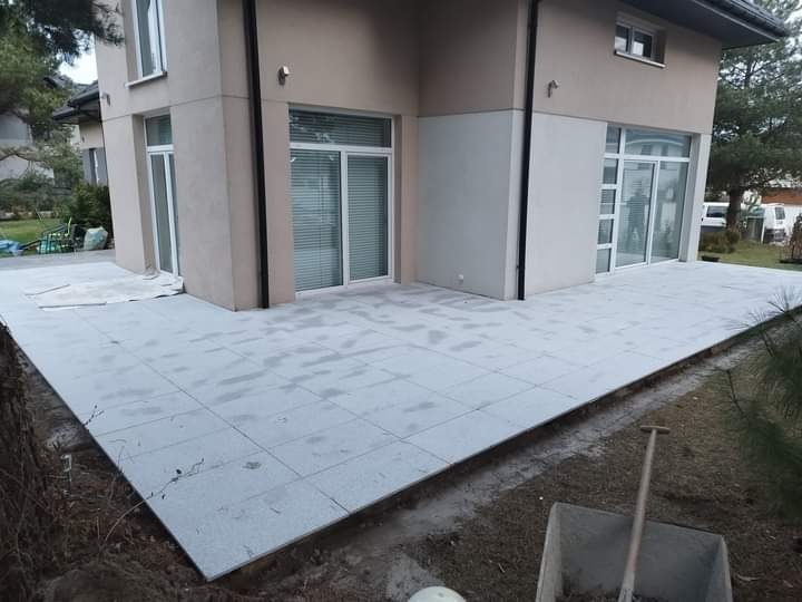 Nowoczesny taras z jasnoszarej kostki ułożonej przy budynku z białymi oknami, widoczny fragment wykopu i narzędzie budowlane.