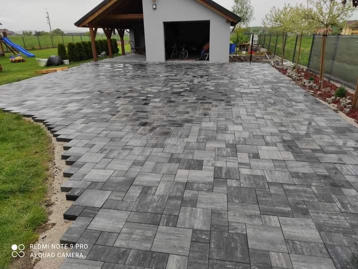 Ułożona szara kostka brukowa w nieregularny wzór przed garażem i trawnikiem. Widoczne niedokończone obrzeża z podsypką piaskową.