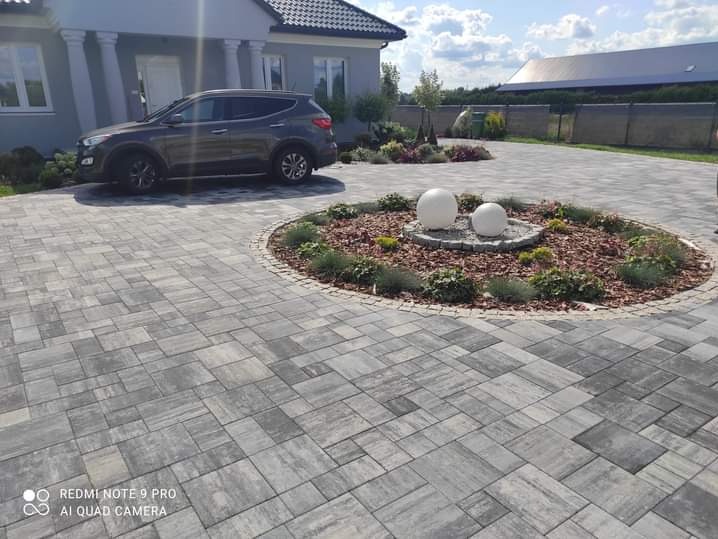 Szara kostka brukowa ułożona na podjeździe do domu jednorodzinnego z centralnie umieszczonym okrągłym kwietnikiem z dwoma białymi kulami dekoracyjnymi, na tle zaparkowanego samochodu i budynku.