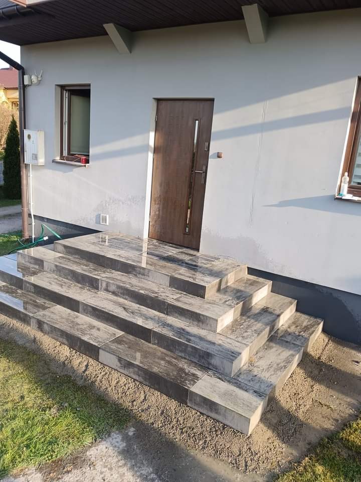 Nowe, czterostopniowe schody wejściowe z szarej kostki brukowej ułożone przed domem z szarą elewacją i brązowymi drzwiami, teren wokół wysypany piaskiem.