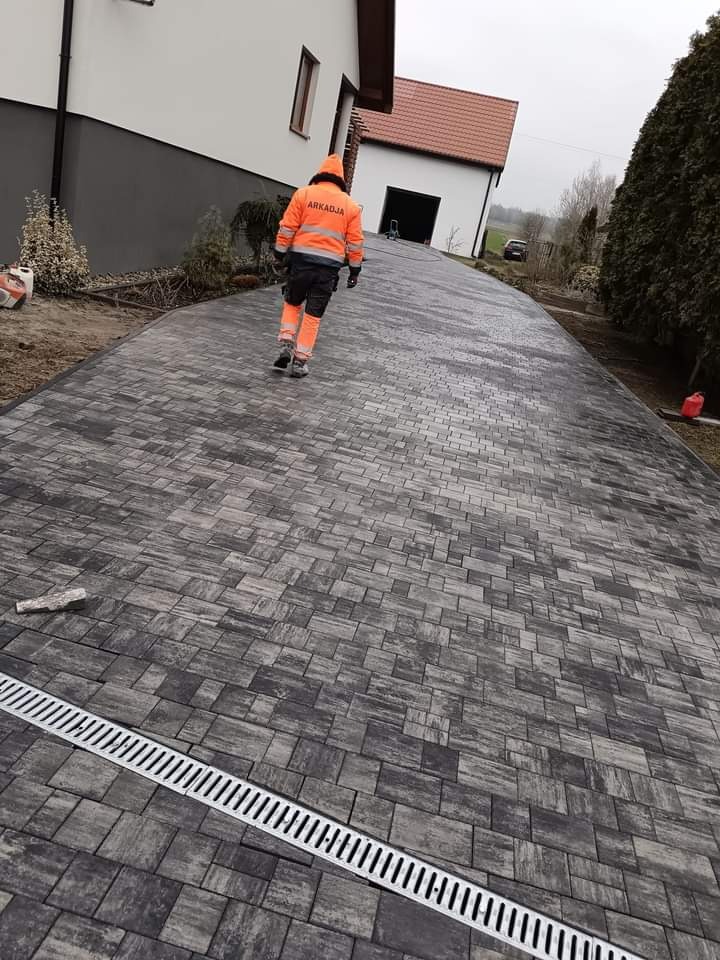 Ukończony podjazd z kostki brukowej w odcieniach szarości, z zamontowanym odpływem liniowym. Pracownik w odblaskowym ubraniu z logo firmy idzie w stronę garażu.