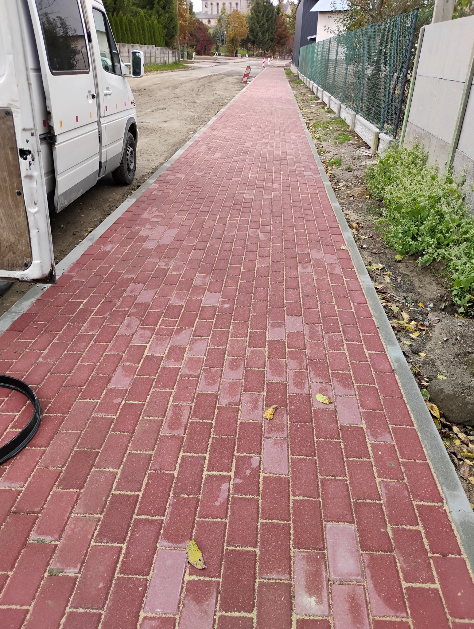 Nowa, długa ścieżka z czerwonej kostki brukowej, z widocznym samochodem dostawczym z otwartymi drzwiami po lewej stronie i zielonym ogrodzeniem po prawej, z kilkoma opadłymi liśćmi na powierzchni.