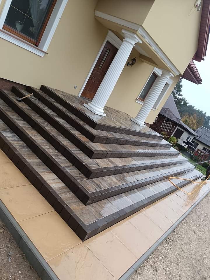 Schody wejściowe do domu wyłożone kostką brukową w odcieniach brązu i beżu, z widocznym młotkiem brukarskim na górnym stopniu, kolumny dekoracyjne przy wejściu.