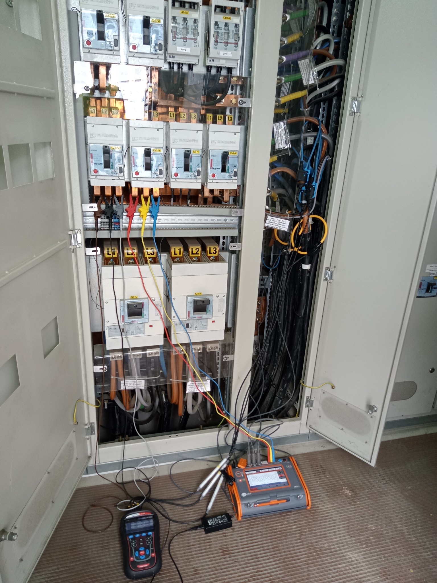 Otwarta szafa elektryczna z widocznymi bezpiecznikami, przewodami i urządzeniami pomiarowymi. Widoczne kable w kolorach: czarnym, niebieskim, żółtym i czerwonym.