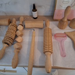 Mind & Body - Studio masażu Kobido - Zestaw drewnianych narzędzi do masażu maderoterapii, w tym wałki o różnych kształtach i skrobak, ułożone na białej serwetce obok buteleczki z olejkiem i grafiki w ramce.