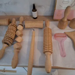 Zestaw drewnianych narzędzi do masażu maderoterapii, w tym wałki o różnych kształtach i skrobak, ułożone na białej serwetce obok buteleczki z olejkiem i grafiki w ramce.