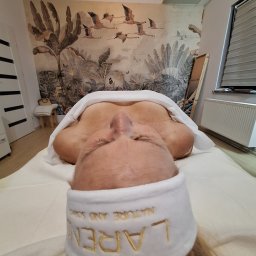 Mind & Body - Studio masażu Kobido - Klientka leżąca na leżance, przykryta białym ręcznikiem, z opaską na włosach z napisem. W tle tapeta z motywami roślinnymi i ptaków.