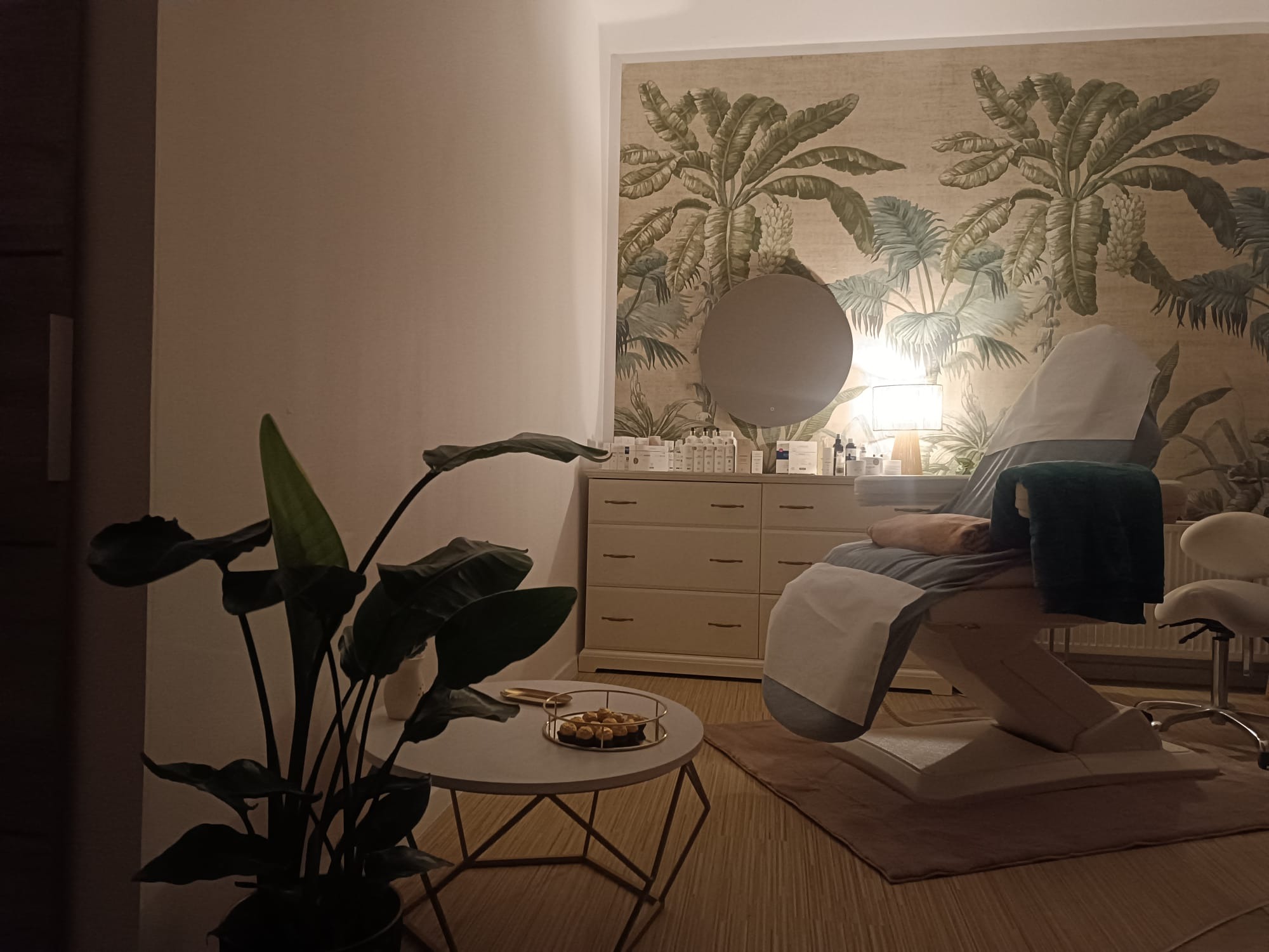 Przytulne wnętrze gabinetu kosmetycznego z fotelem zabiegowym przykrytym pokrowcem, komodą z kosmetykami, okrągłym lustrem na ścianie z tapetą w motywy tropikalne oraz stolikiem kawowym...