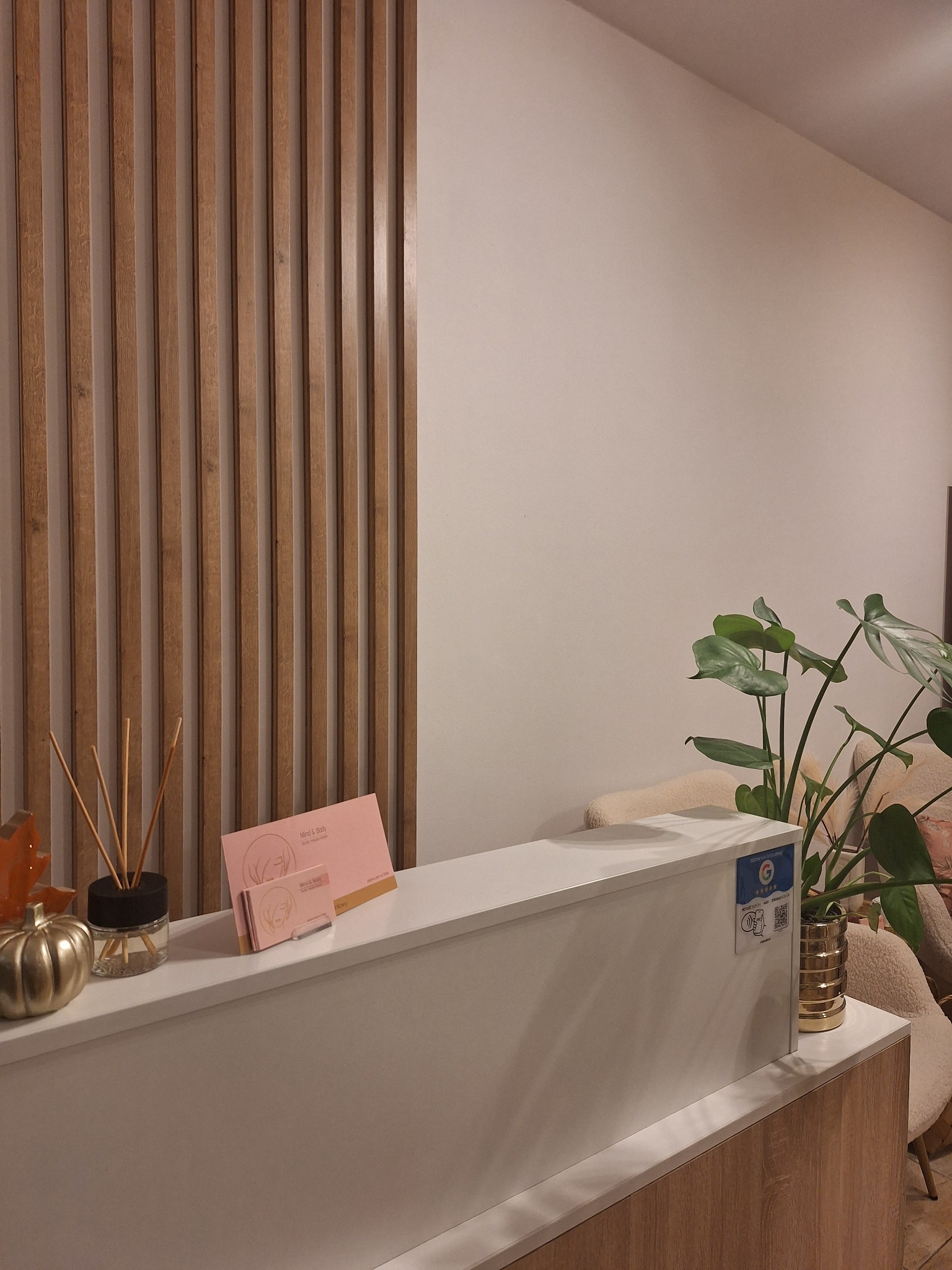 Recepcja w jasnych barwach z drewnianymi elementami dekoracyjnymi, dyfuzorem zapachowym, złotą dynią i ulotkami na blacie, oraz rośliną doniczkową w tle.