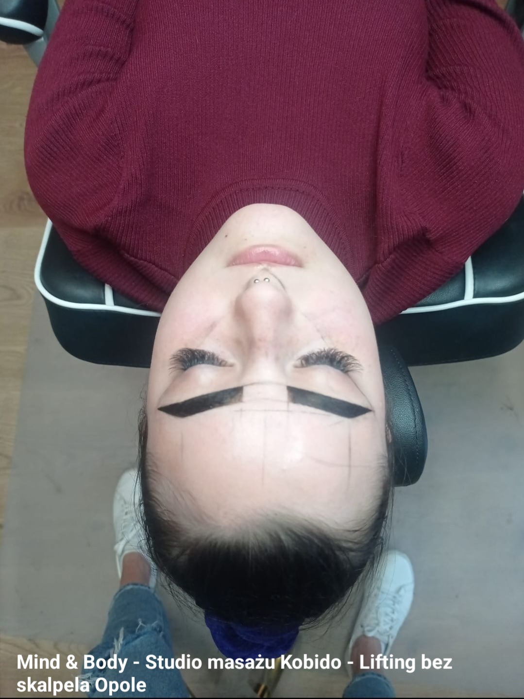 Klientka w trakcie zabiegu stylizacji brwi z nałożonymi ciemnymi paskami henny i precyzyjnymi liniami pomiarowymi, widok z góry, w tle fotel kosmetyczny i fragment butów stylistki.