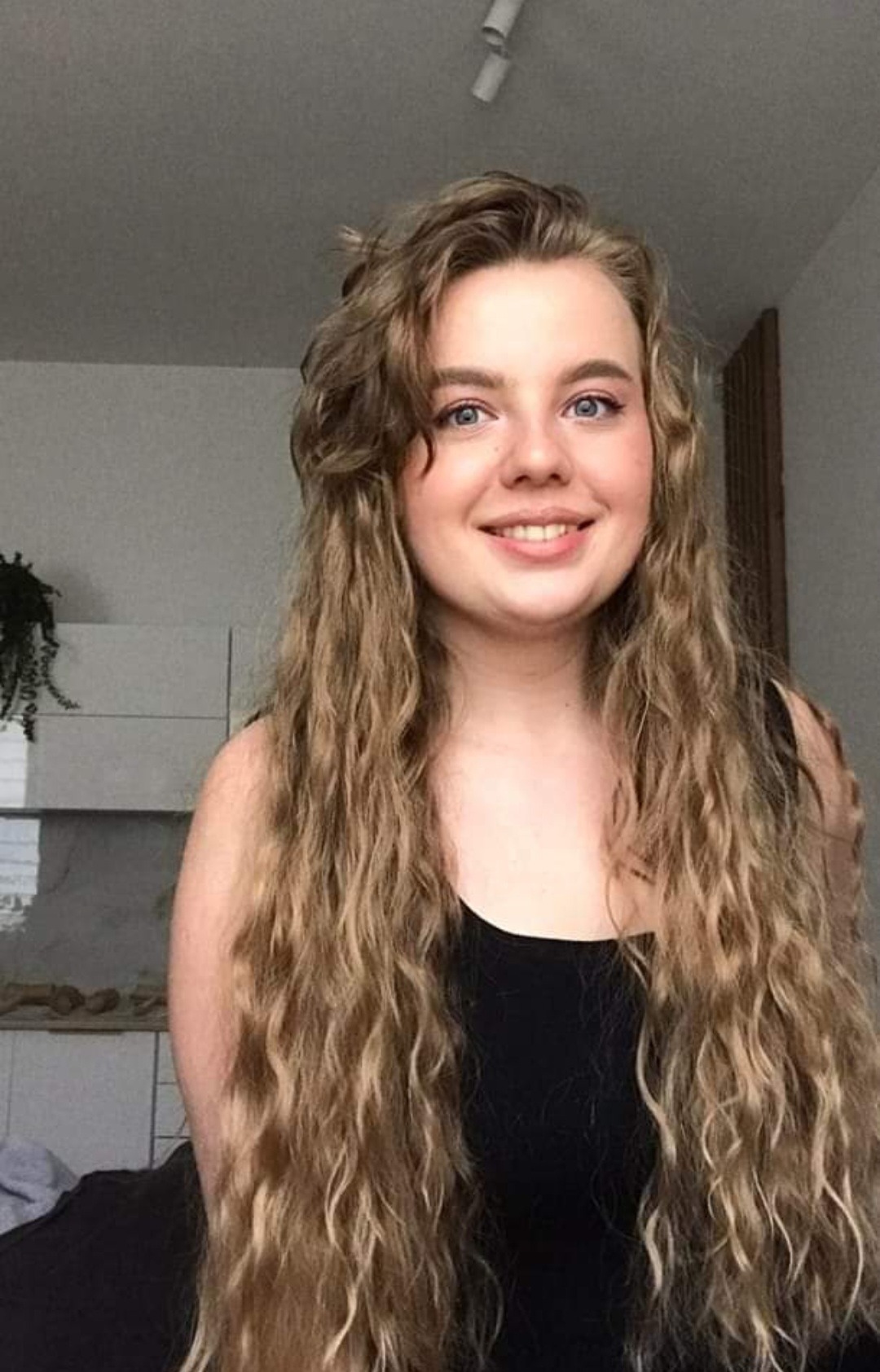 Portret uśmiechniętej blondynki o niebieskich oczach i długich, kręconych włosach w czarnym topie, na tle jasnego wnętrza z elementami dekoracyjnymi.