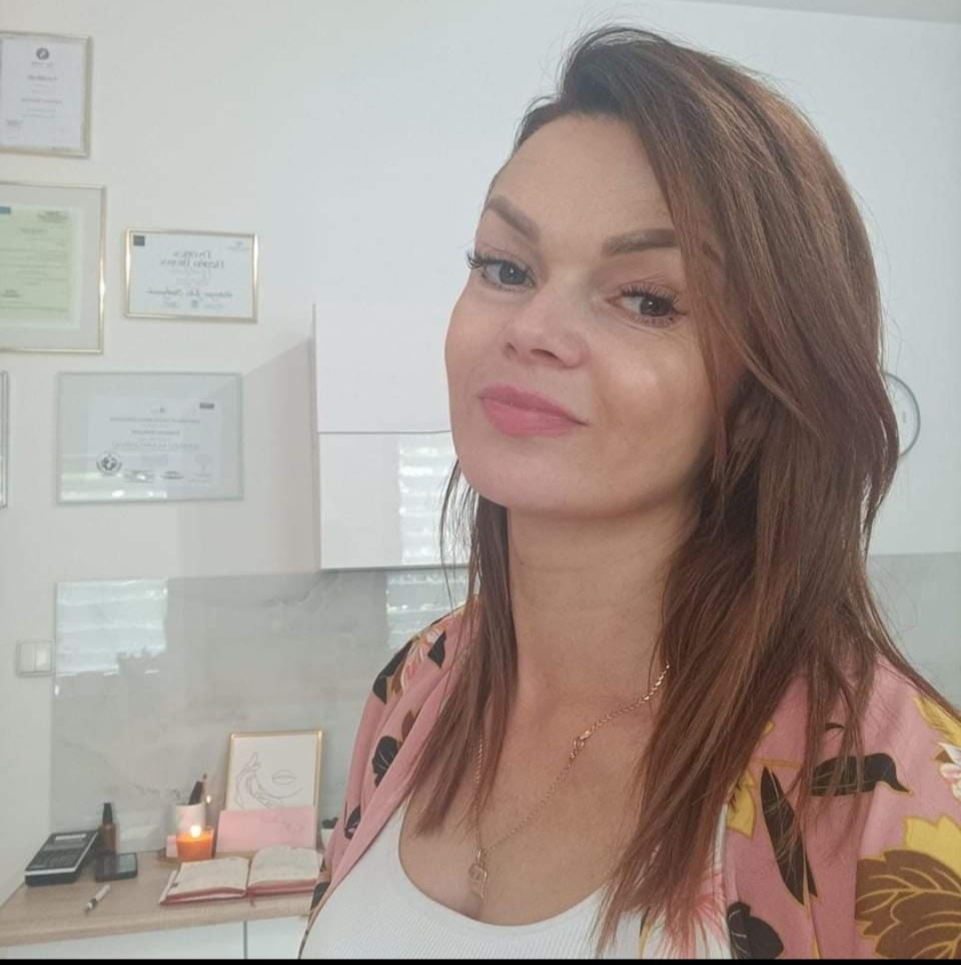 Portret kobiety z brązowymi włosami, delikatnym makijażem i złotym naszyjnikiem na tle gabinetu z certyfikatami na ścianie i biurkiem z notatnikiem, świecą i kalkulatorem.