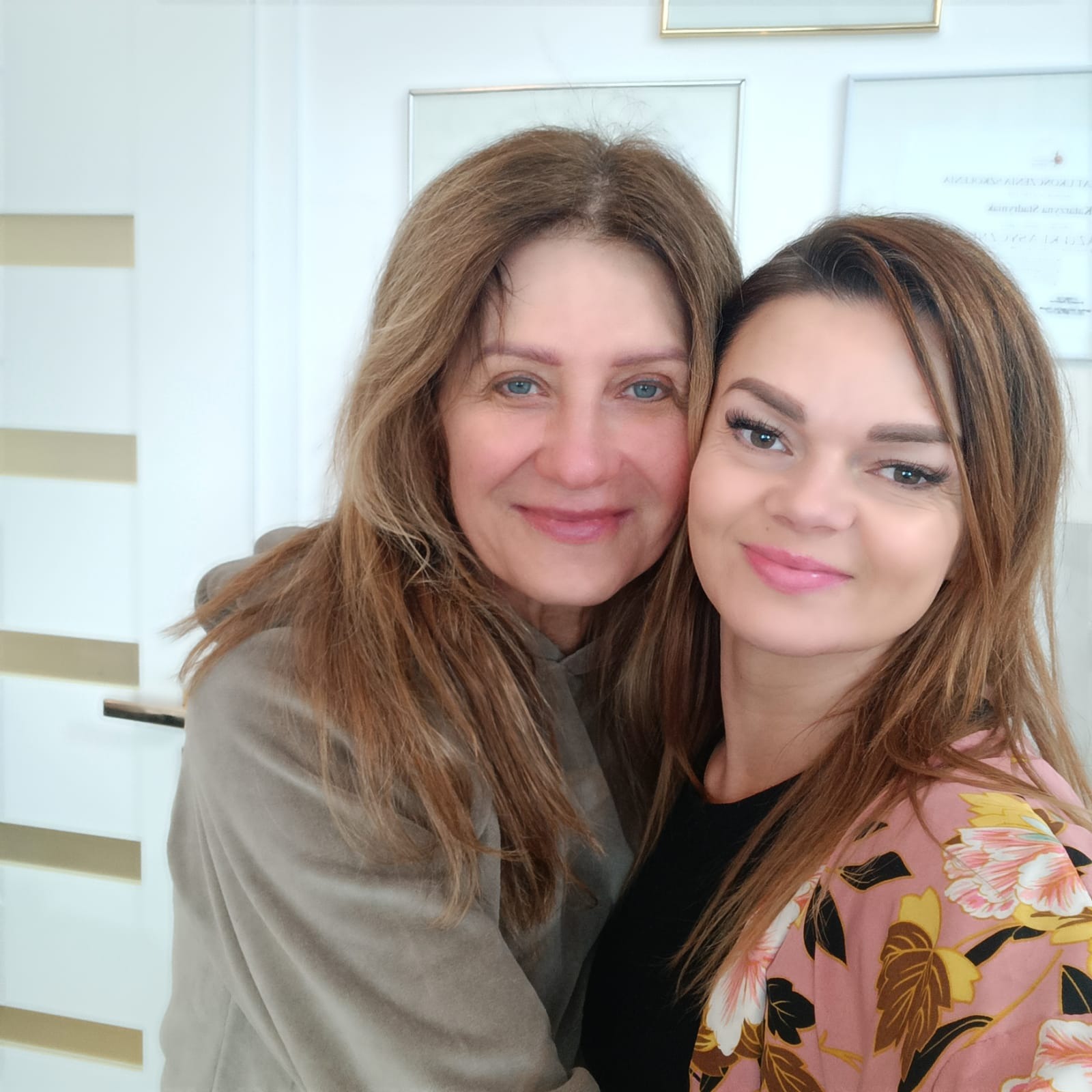 Portret uśmiechniętych kobiet, jedna z widocznymi, świeżo wystylizowanymi brwiami i rzęsami, w tle ściana z ramkami.