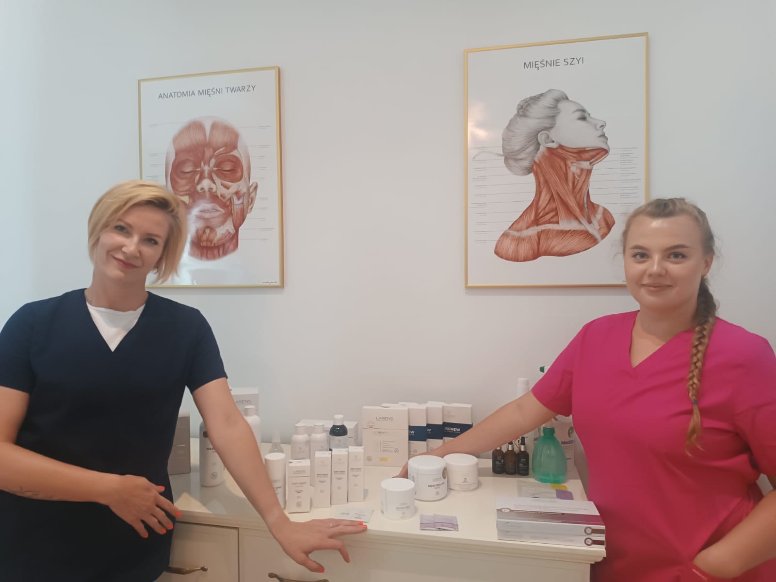 Dwie kobiety w strojach roboczych pozują w gabinecie z plakatami anatomicznymi i kosmetykami marki Larens i Renew na komodzie.