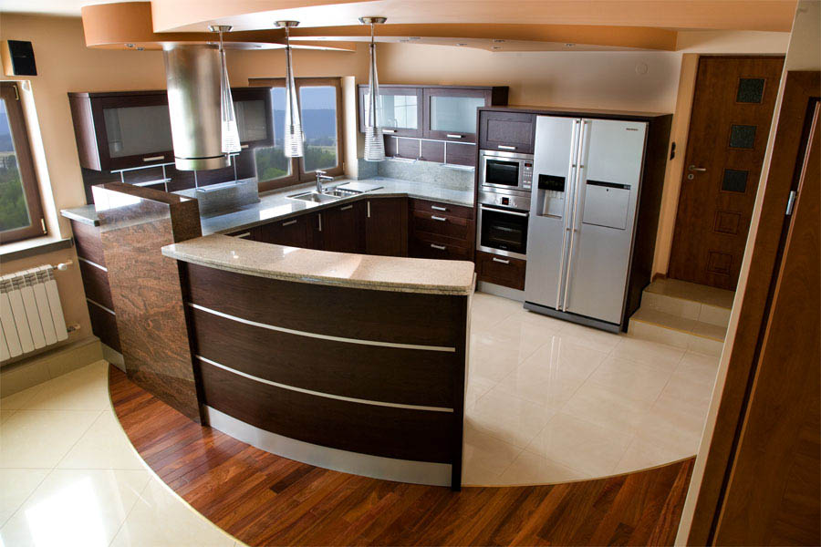 Nowoczesna kuchnia z ciemnymi drewnianymi szafkami, blatem z jasnego granitu i półwyspem z zaokrąglonym frontem, oświetlona wiszącymi lampami i wpadającym światłem z okna.