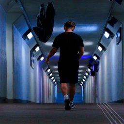 Foto Art-Live - Mężczyzna idący przez oświetlony tunel, rzucający czarną bluzę w powietrze, widok z tyłu, z akcentem na dynamiczne ujęcie bluzy w locie.