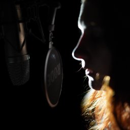 Foto Art-Live - Profil kobiety z rudymi włosami śpiewającej do mikrofonu studyjnego z pop filtrem w ciemnym pomieszczeniu.