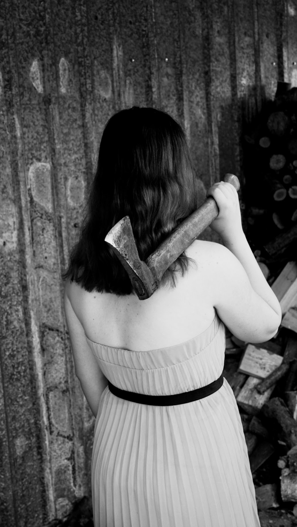 Czarno-białe zdjęcie kobiety w sukience bez ramiączek, trzymającej na ramieniu topór przed stertą drewna i blaszanej ściany.
