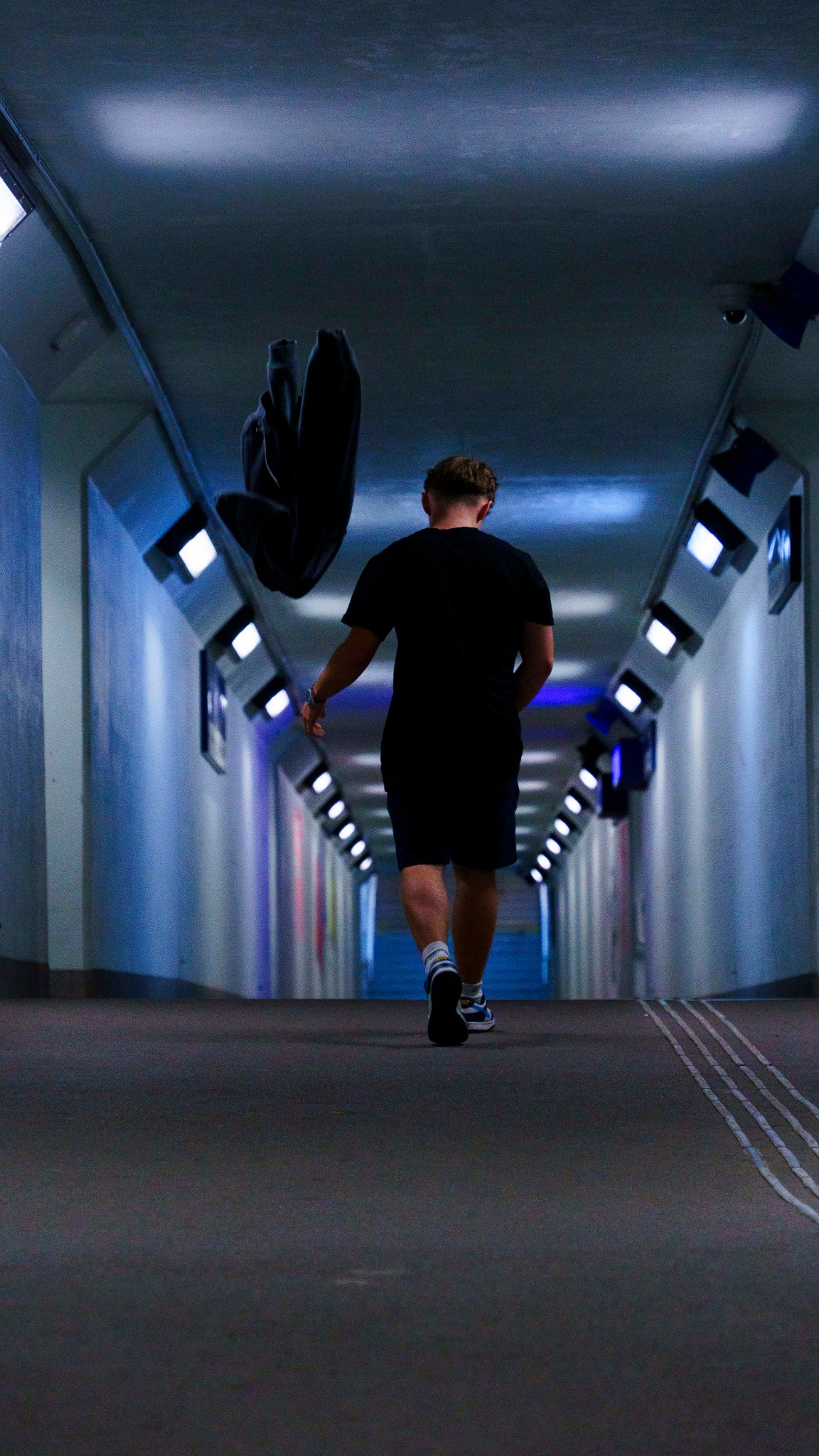 Mężczyzna idący przez oświetlony tunel, rzucający czarną bluzę w powietrze, widok z tyłu, z akcentem na dynamiczne ujęcie bluzy w locie.