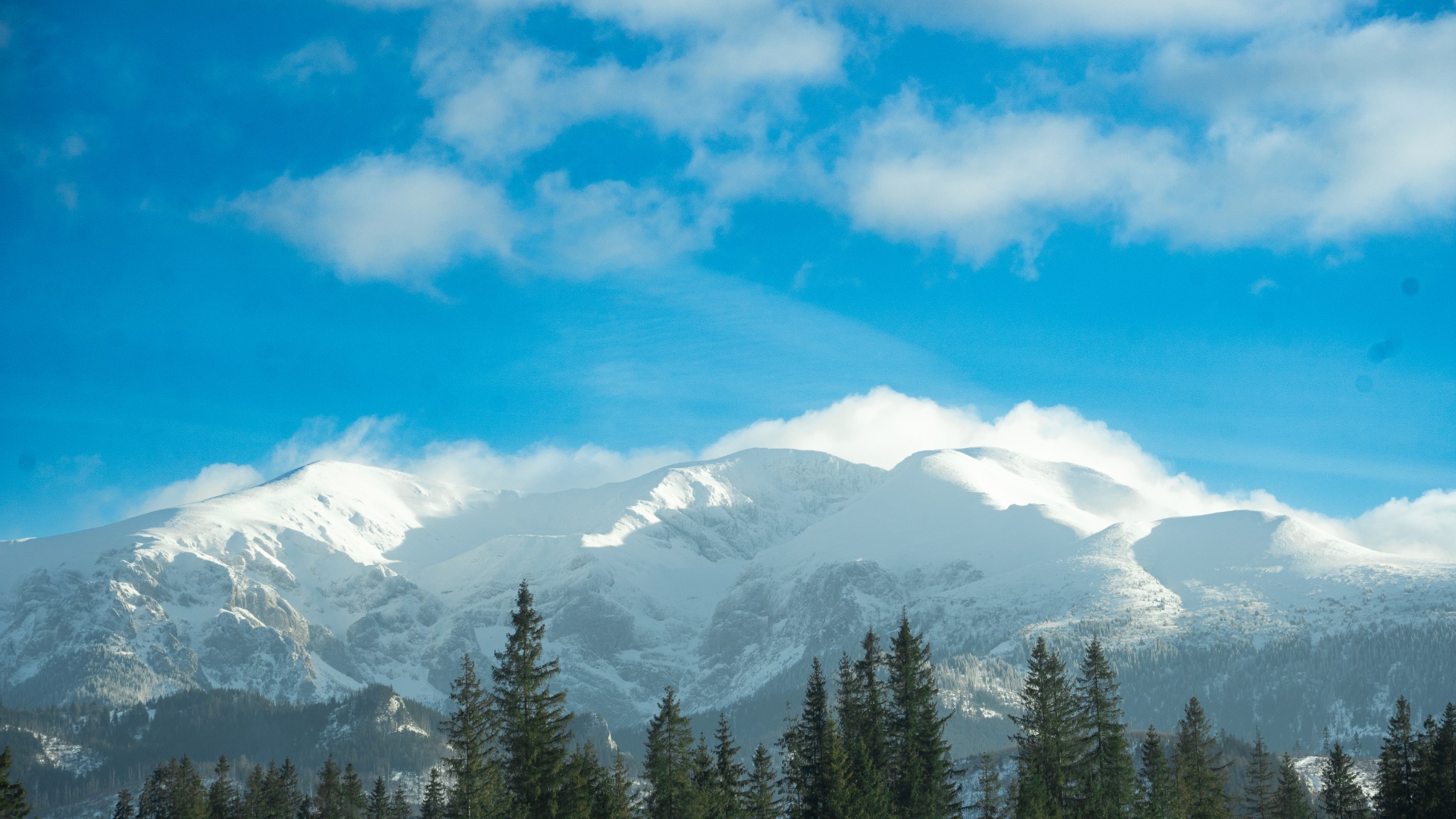 Panoramiczny widok na ośnieżone szczyty górskie z lasem iglastym u podnóża, błękitne niebo z białymi obłokami w tle.