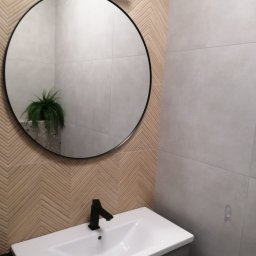 Michał Domański - Nowoczesna łazienka z okrągłym lustrem i umywalką na szafce. Na ścianie płytki w jodełkę i szare płytki. W lustrze odbija się roślina doniczkowa.