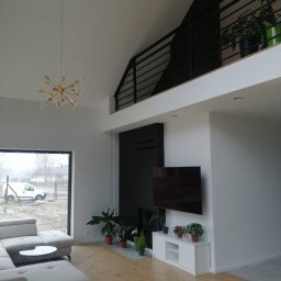 Michał Domański - Jasne wnętrze domu z antresolą i dużym oknem. Widoczny narożnik, stolik kawowy, szafka RTV z roślinami i telewizor. Na antresoli balustrada i donice z zielenią.