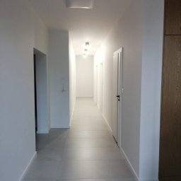 Michał Domański - Długi korytarz z szarymi płytkami podłogowymi i białymi ścianami. Widoczne drzwi i zabudowa szafy w odcieniach drewna. Minimalistyczne oświetlenie sufitowe.