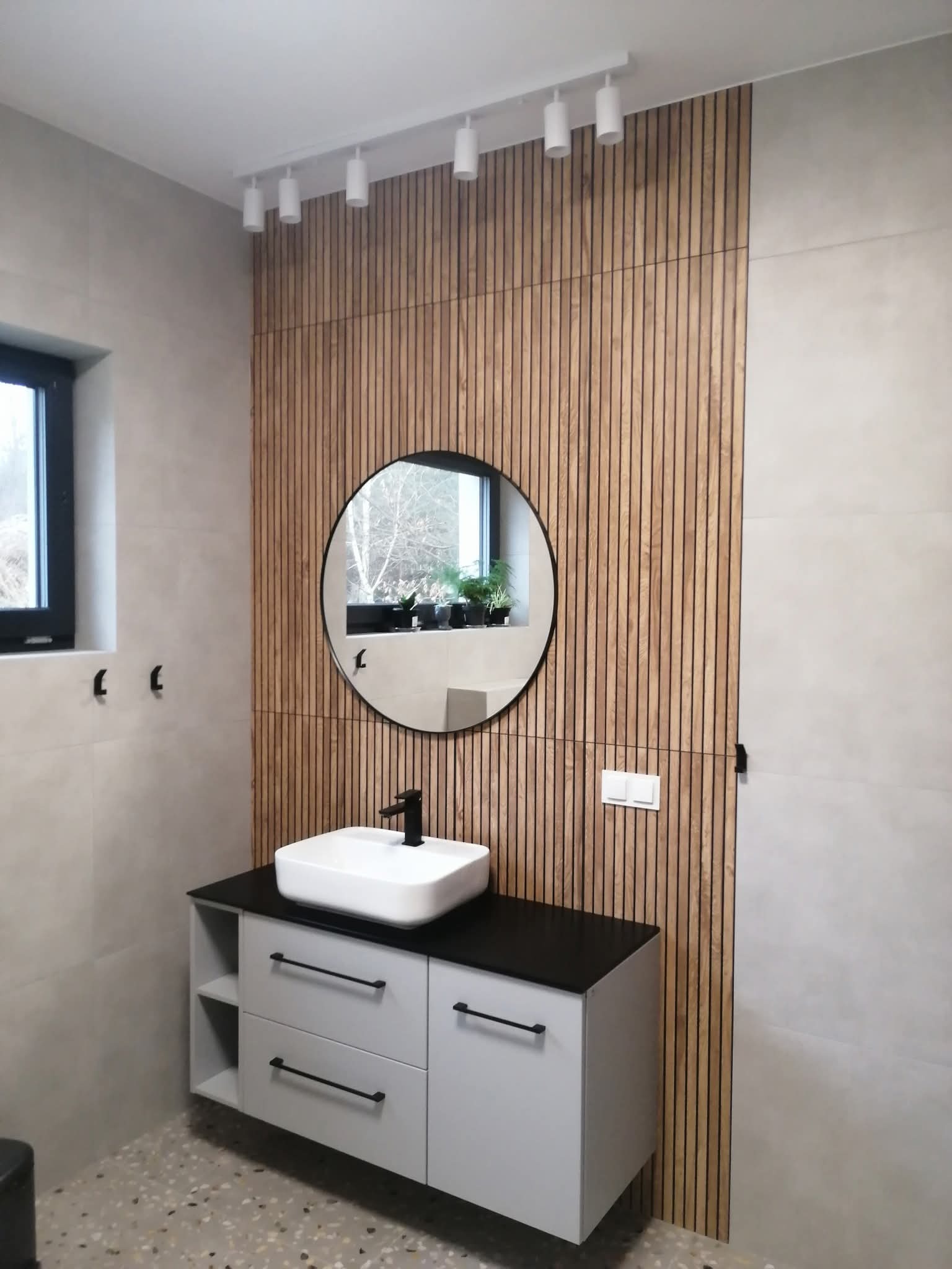 Nowoczesna łazienka z drewnianą ścianą, okrągłym lustrem i białą umywalką na szafce z szufladami. Oświetlenie punktowe na suficie. Minimalistyczny design.