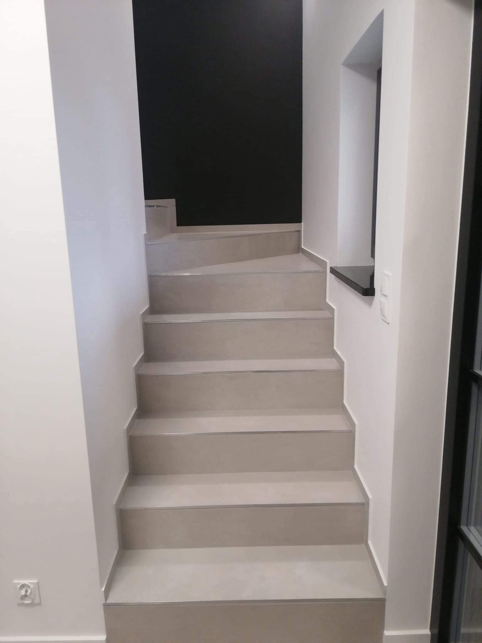 Wykończone schody wewnętrzne z szarymi płytkami, białe ściany i czarny akcent kolorystyczny na ścianie w tle. Minimalistyczny design z oknem po prawej stronie.