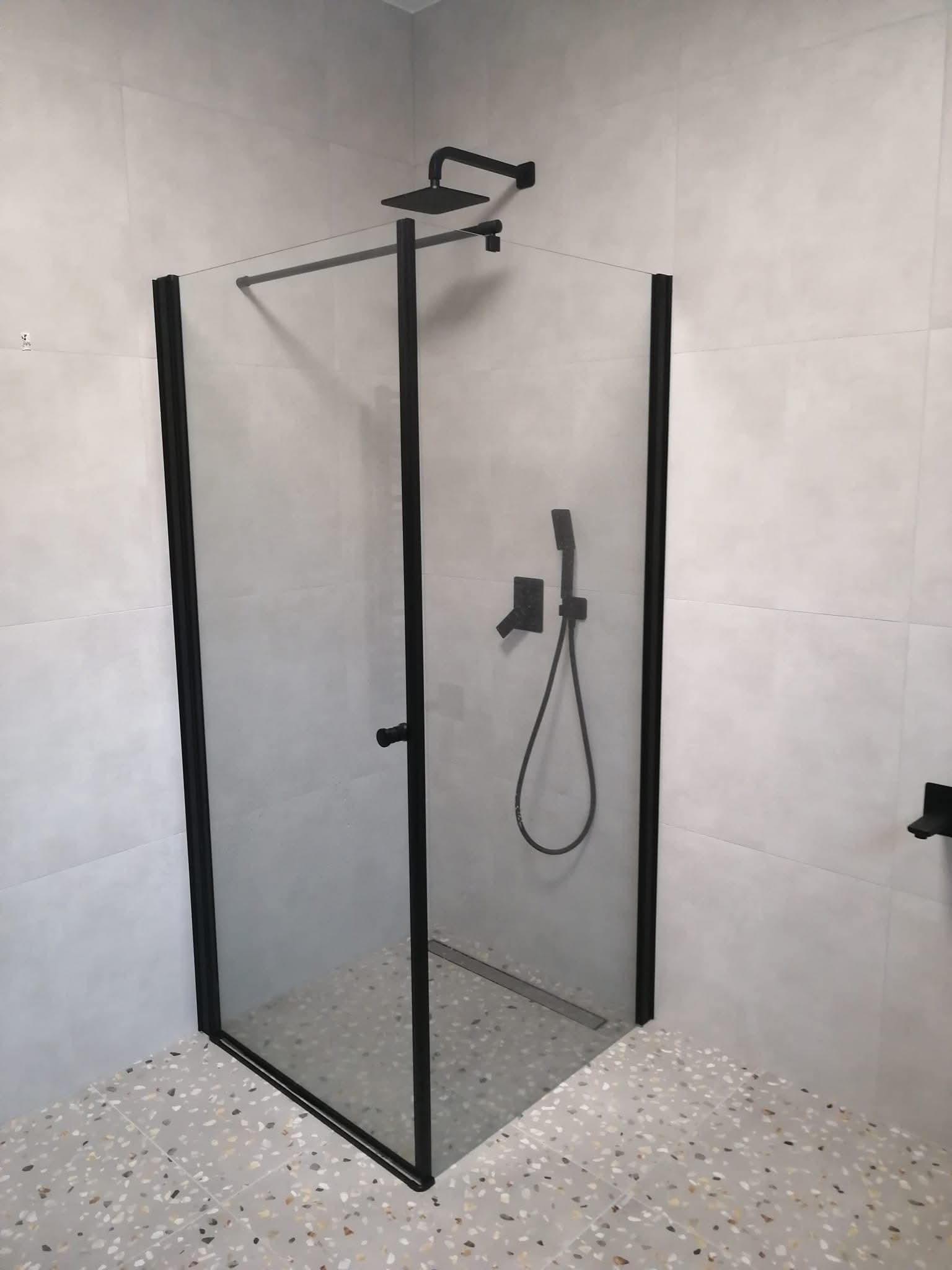 Nowoczesna kabina prysznicowa z czarnymi profilami na tle jasnych płytek i podłogi lastryko. Minimalistyczny design, armatura w kolorze czarnym.