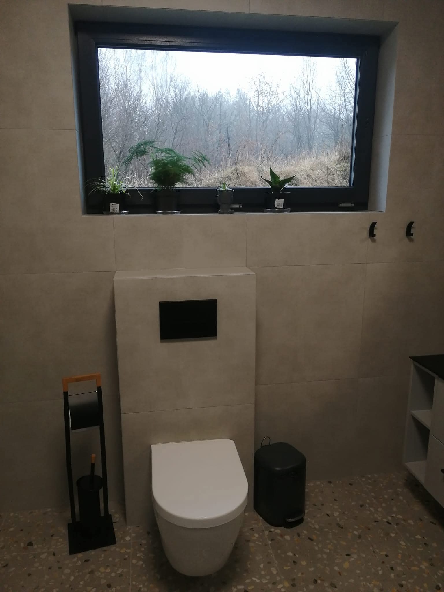 Nowoczesna łazienka z beżowymi płytkami, wiszącą toaletą i oknem z widokiem na drzewa. Na parapecie doniczki z roślinami. Minimalistyczny design.