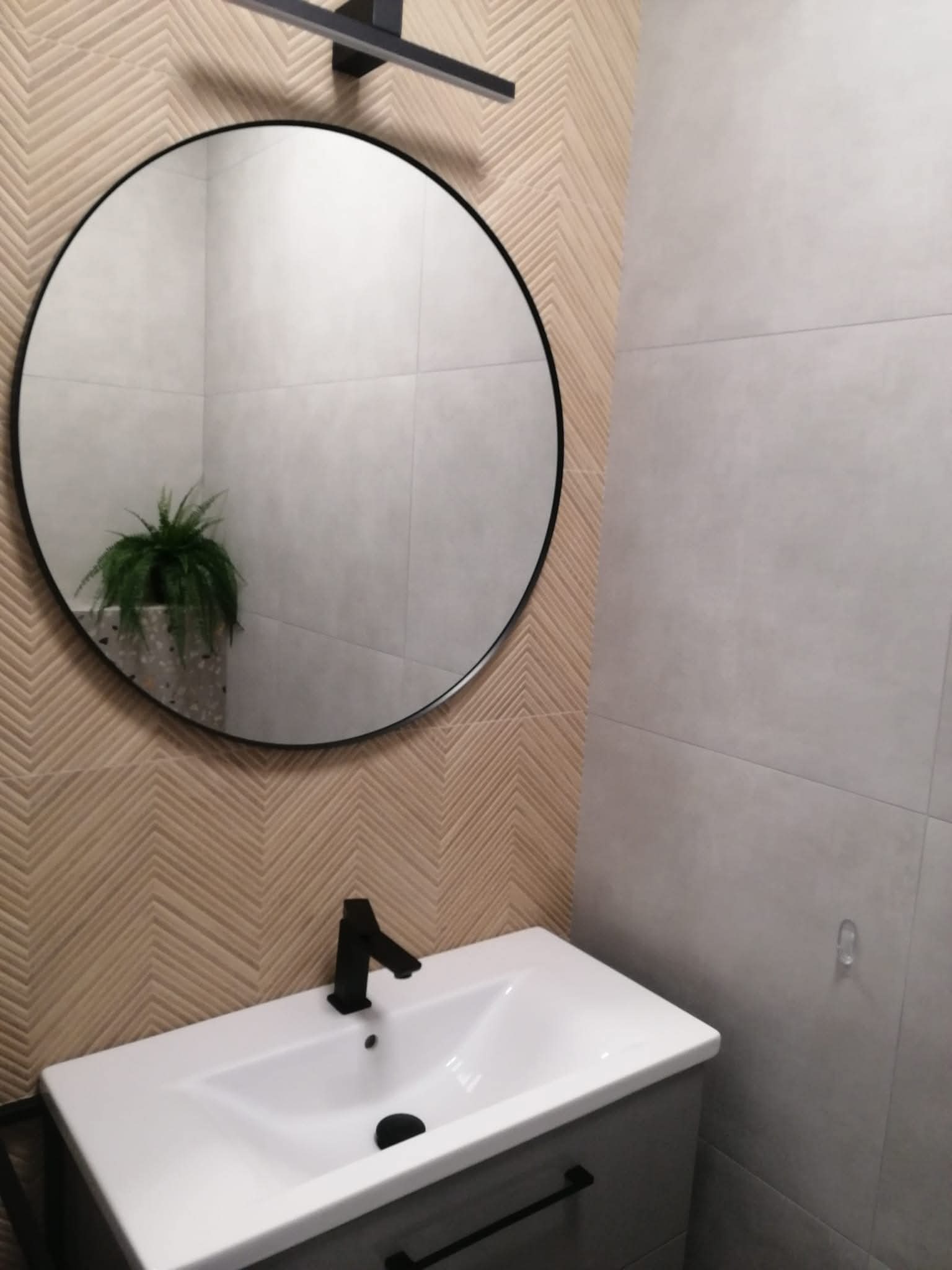 Nowoczesna łazienka z okrągłym lustrem i umywalką na szafce. Na ścianie płytki w jodełkę i szare płytki. W lustrze odbija się roślina doniczkowa.