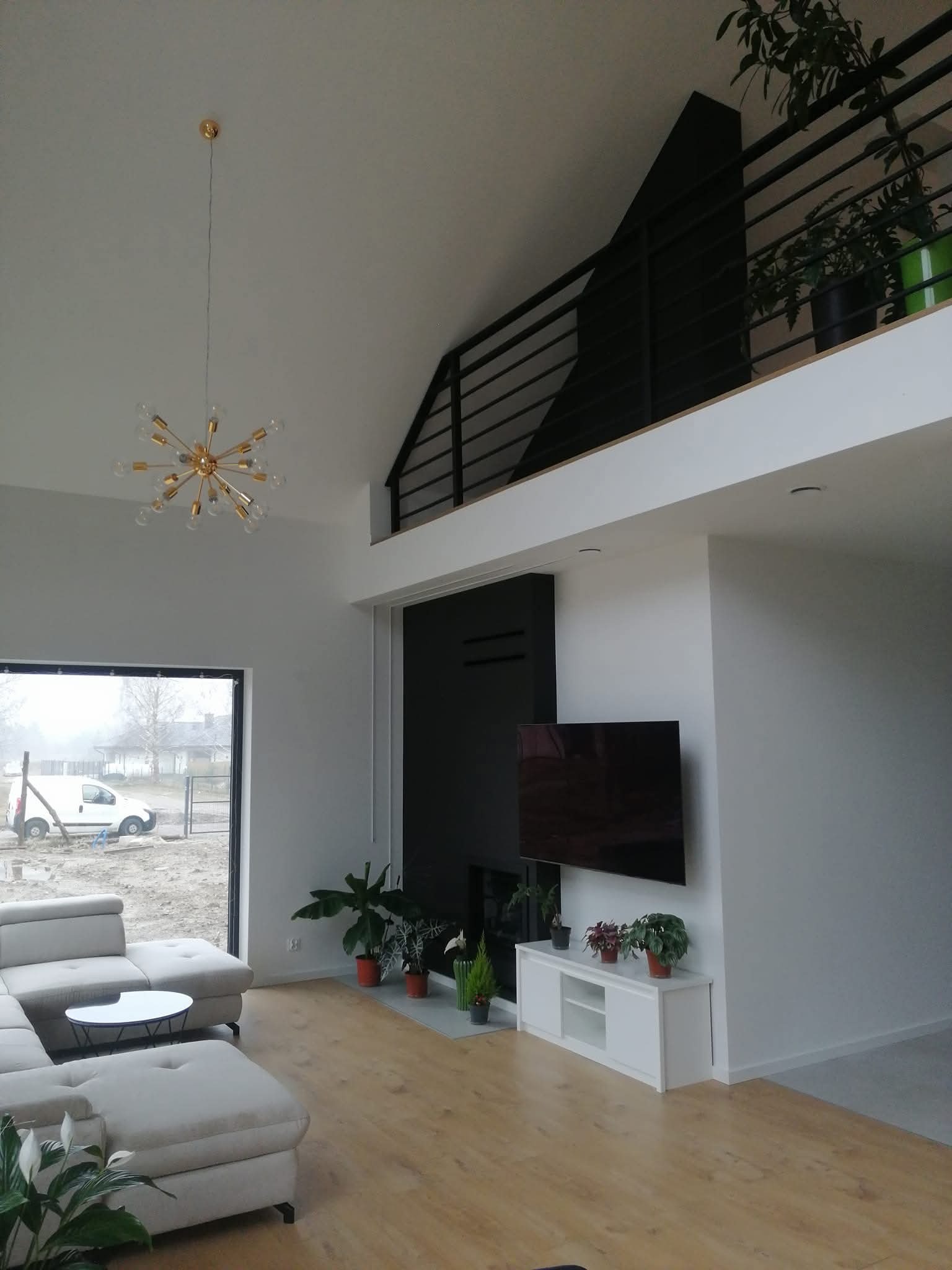 Jasne wnętrze domu z antresolą i dużym oknem. Widoczny narożnik, stolik kawowy, szafka RTV z roślinami i telewizor. Na antresoli balustrada i donice z zielenią.