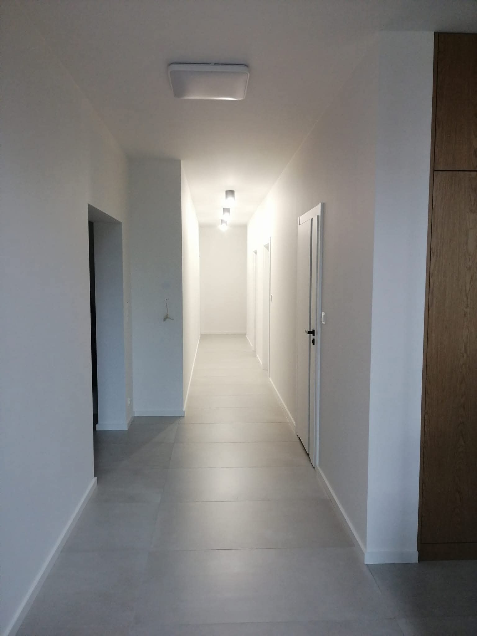 Długi korytarz z szarymi płytkami podłogowymi i białymi ścianami. Widoczne drzwi i zabudowa szafy w odcieniach drewna. Minimalistyczne oświetlenie sufitowe.