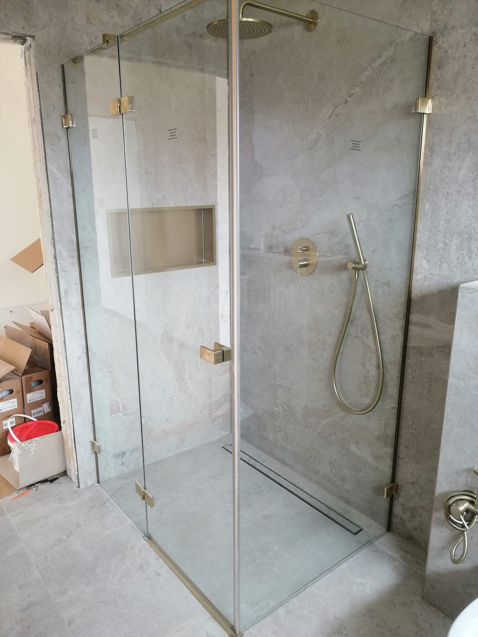 Szklana kabina prysznicowa z złotymi okuciami, w łazience wykończonej płytami imitującymi marmur. Widoczna wnęka w ścianie i armatura w kolorze złota.