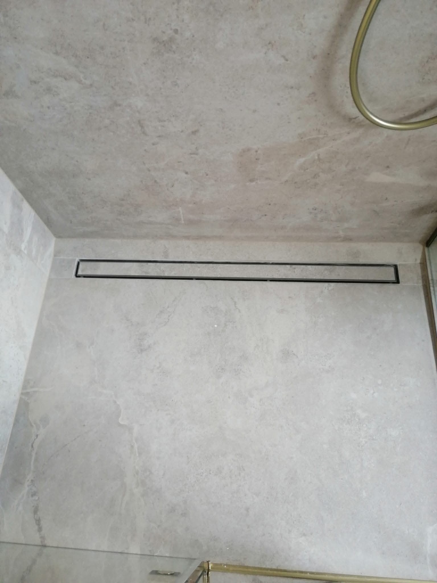 Minimalistyczna łazienka z odpływem liniowym w podłodze, wykończona płytkami imitującymi marmur. Widok z dołu, złote elementy armatury.