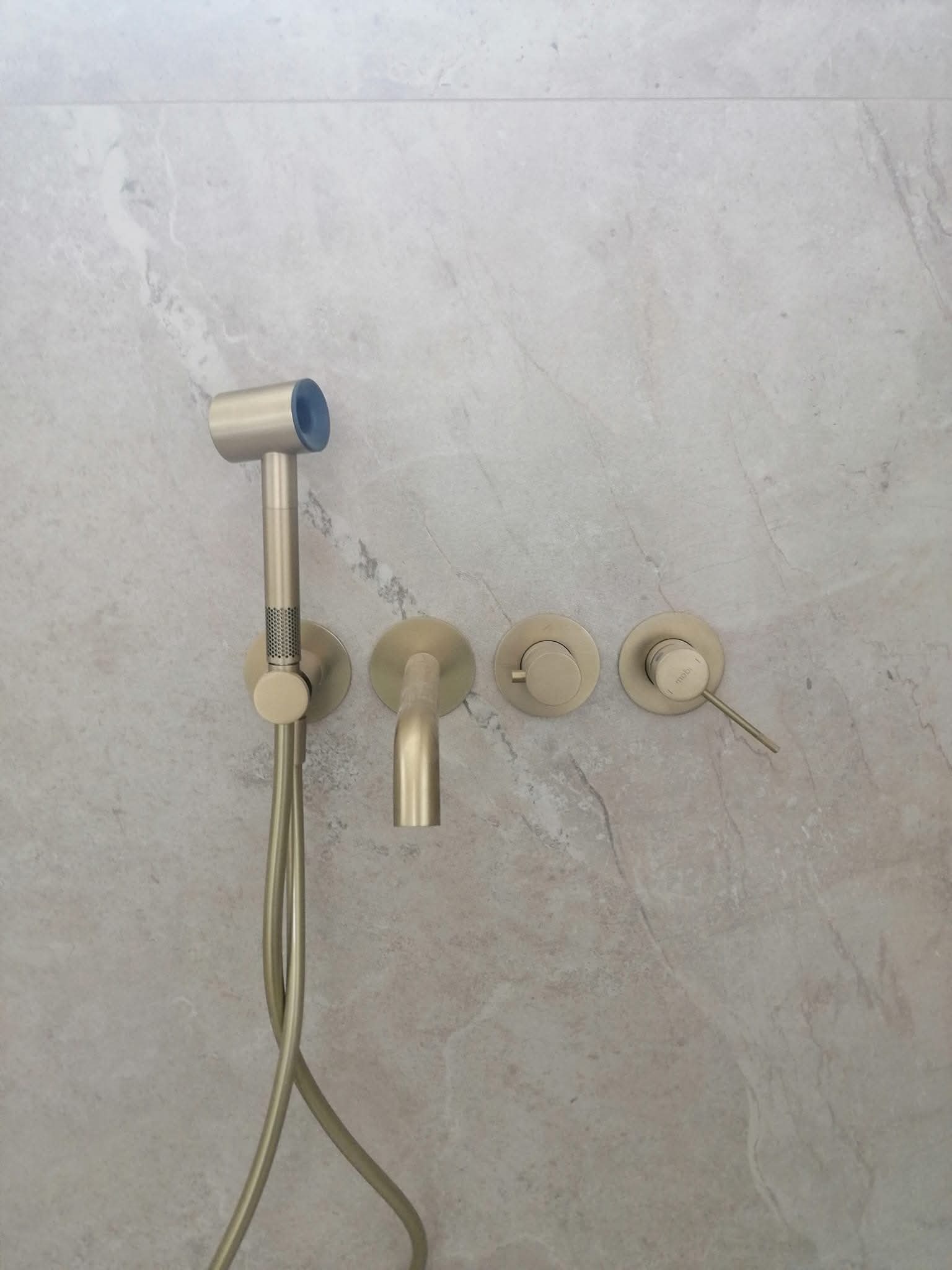 Bidetta w kolorze szczotkowanego złota, zamontowana na szarej, marmurkowej ścianie w łazience. Minimalistyczny design, widoczne elementy instalacji.