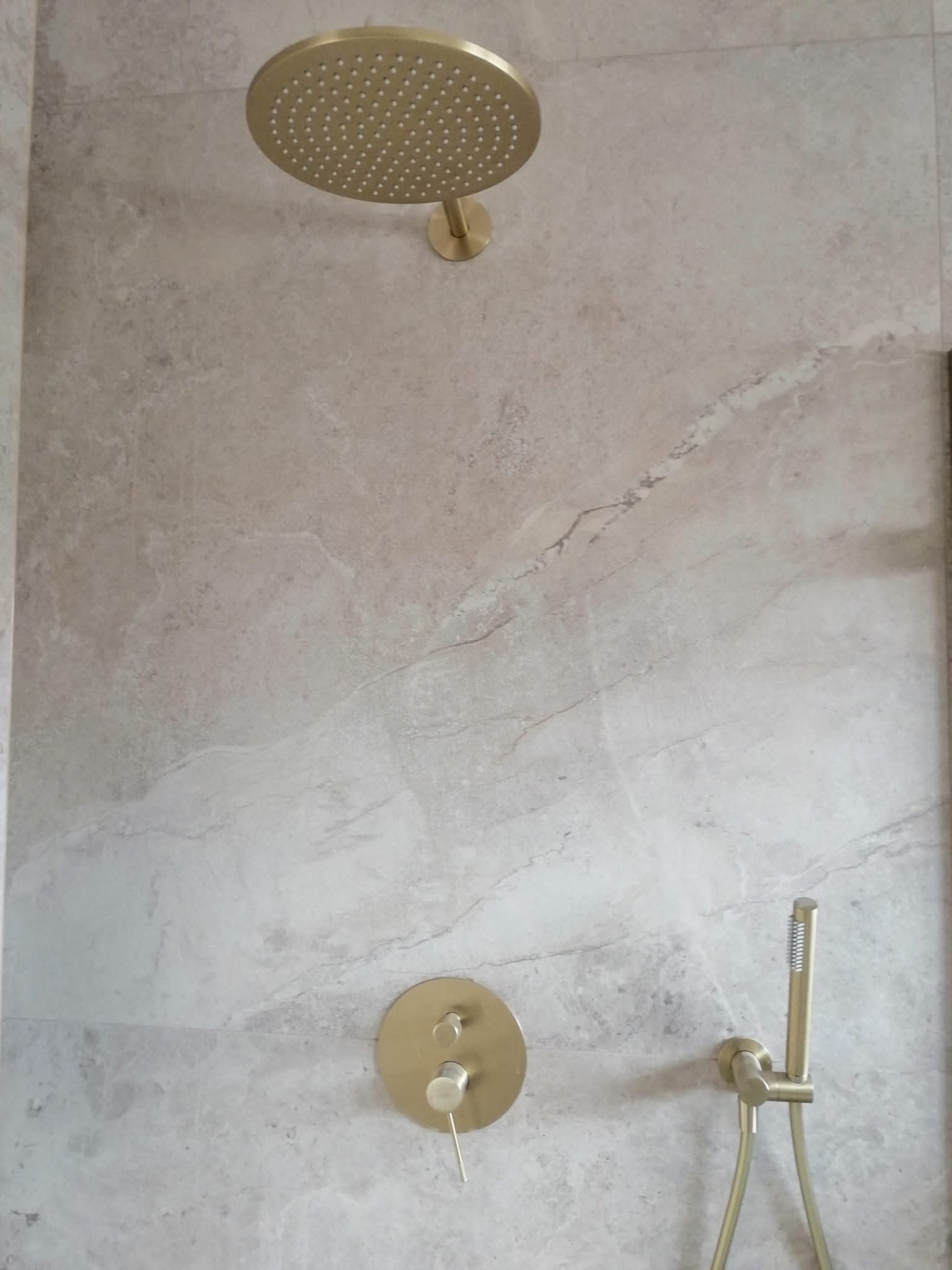 Beżowa ściana z marmurowym wzorem, złota deszczownica, bateria i słuchawka prysznicowa. Minimalistyczny design łazienki z akcentami w kolorze złota.