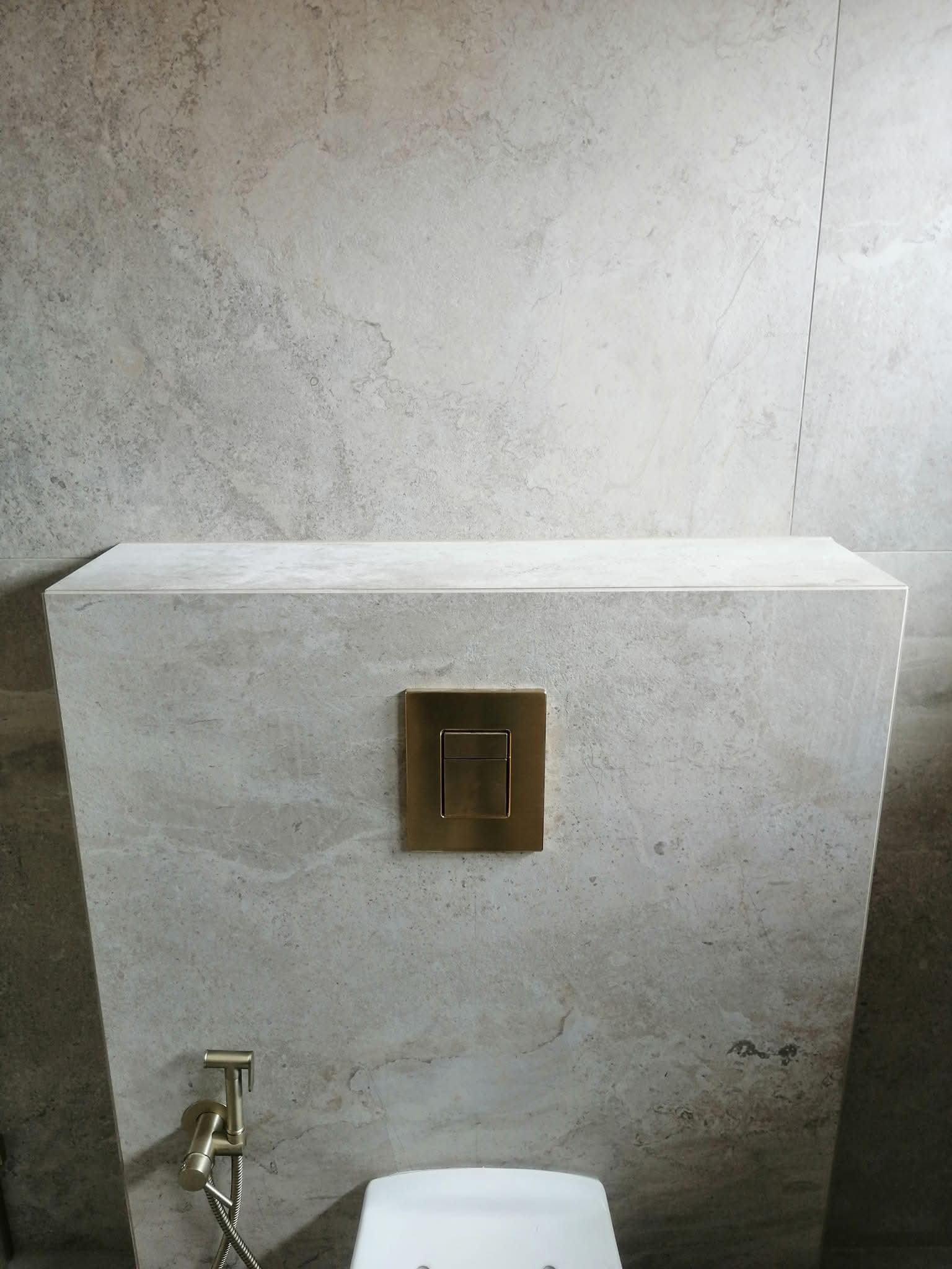 Szara ściana z płytek imitujących kamień, zabudowa toalety z przyciskiem w kolorze złotym i bidetką. Minimalistyczne wykończenie łazienki.