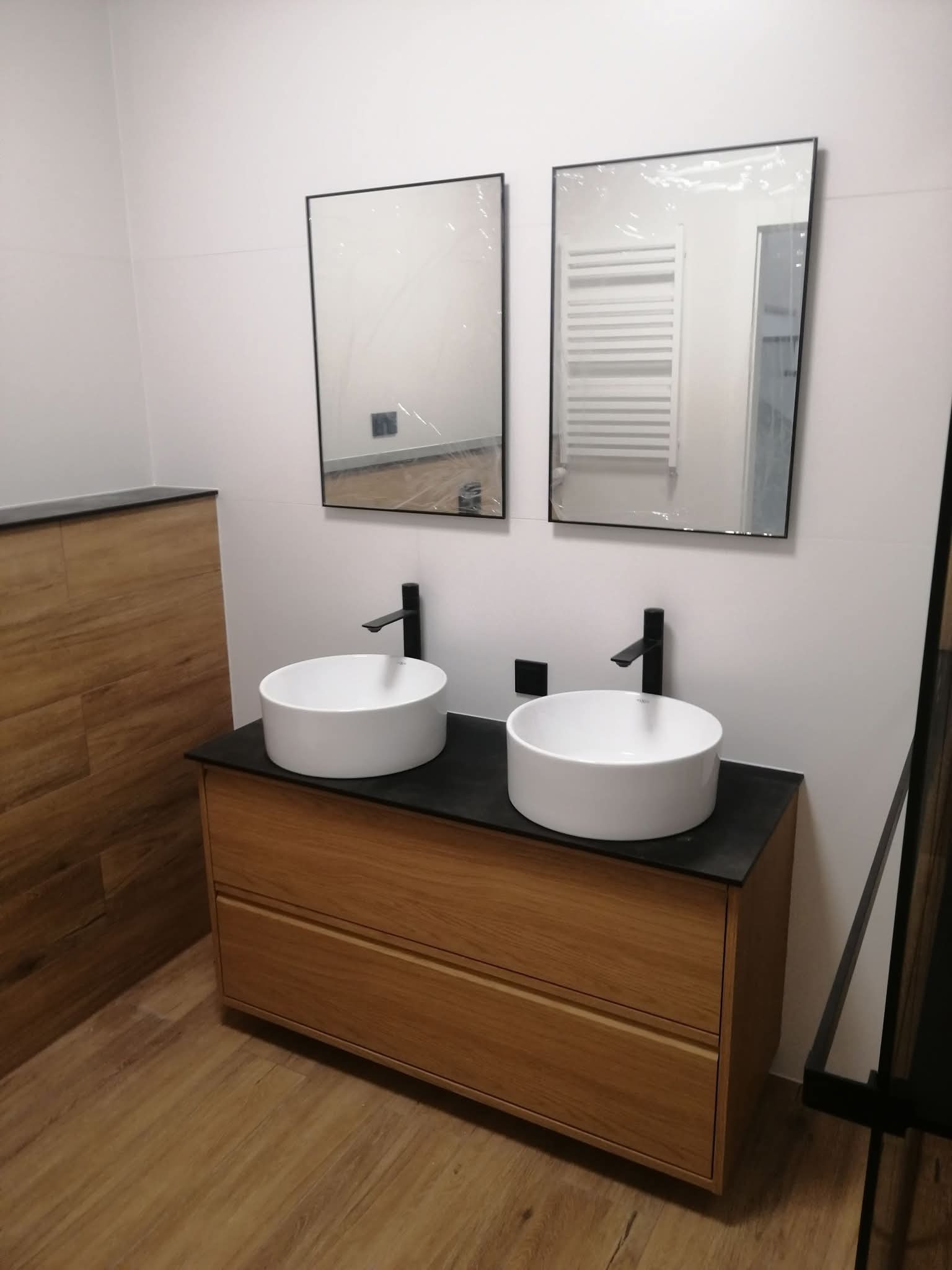 Nowoczesna łazienka z drewnianą szafką pod umywalki, czarnym blatem i armaturą. Dwa lustra w czarnych ramach. Na ścianach białe płytki imitujące drewno.