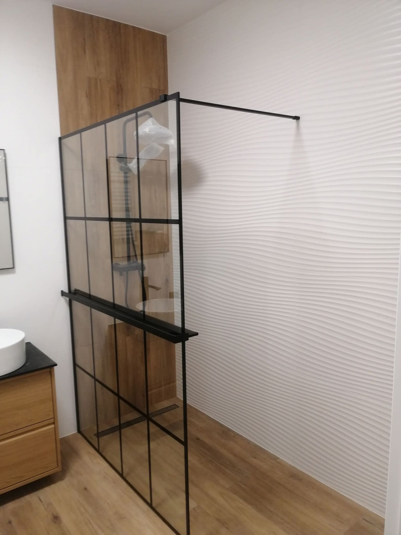 Nowoczesna kabina prysznicowa z czarnymi profilami i szklanymi panelami, drewnopodobne płytki na ścianie i podłodze, dekoracyjna biała ściana z efektem fal.