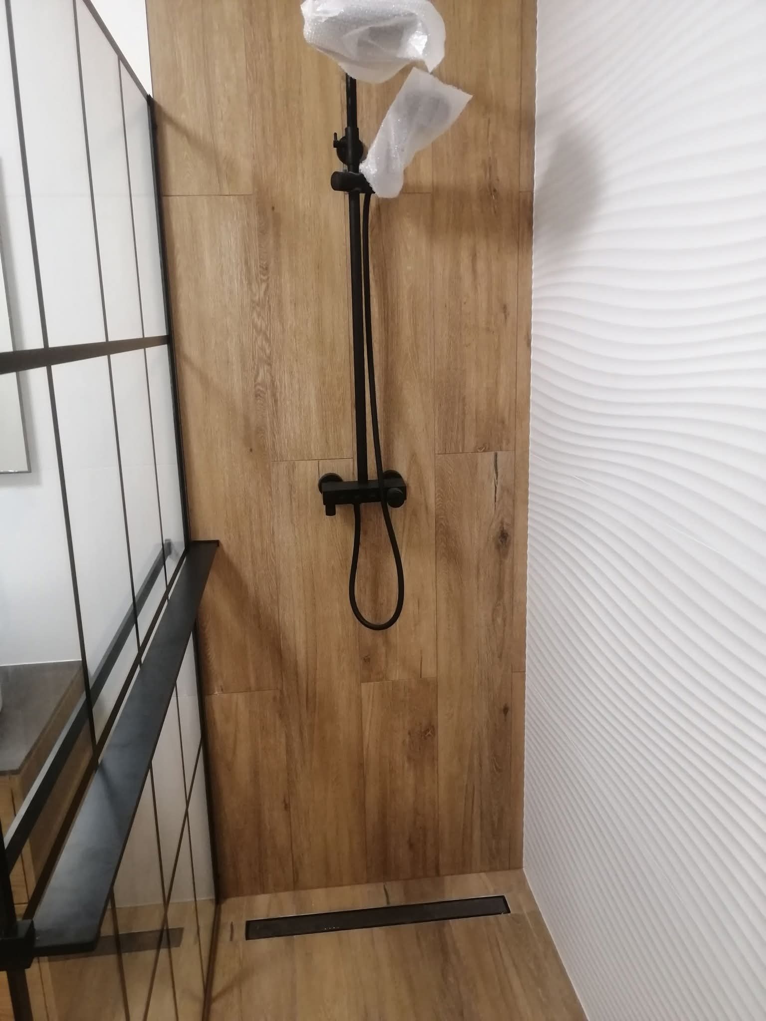 Nowoczesna łazienka z kabiną typu walk-in, wykończona płytkami imitującymi drewno i białymi panelami 3D. Czarna armatura prysznicowa z folią ochronną.