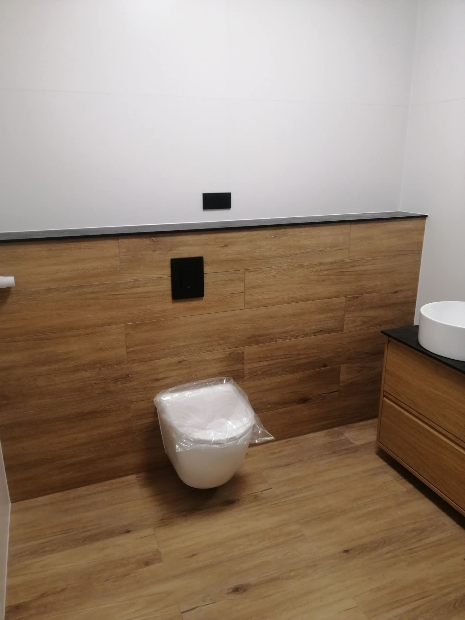 Nowoczesna łazienka z drewnopodobnymi płytkami na ścianie i podłodze, białą umywalką nablatową i podwieszaną toaletą w folii ochronnej. Minimalistyczny design.