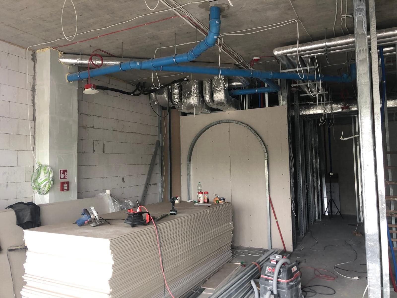 Wnętrze w trakcie prac wykończeniowych: płyty gipsowe, instalacje pod sufitem, metalowe profile. Widoczny etap budowy ścianki działowej z łukowym przejściem.