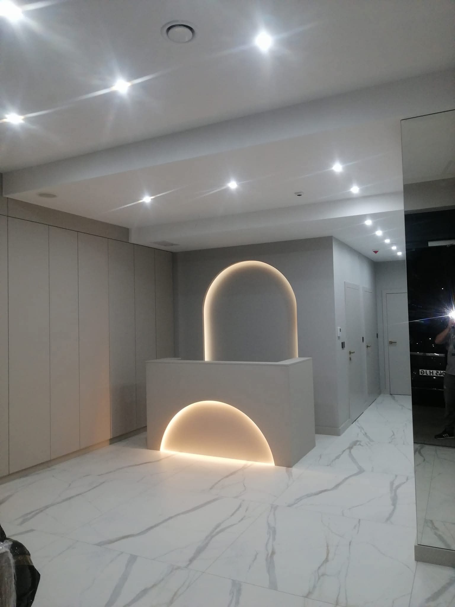 Nowoczesne wnętrze z recepcją oświetloną półkolistym światłem, marmurowa podłoga i minimalistyczne szafy w zabudowie. Widoczne oświetlenie sufitowe.
