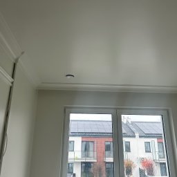 BUD-MAR USŁUGI BUDOWLANE MARCIN MAŃKOWSKI - Wnętrze po remoncie: gładka, biała ściana z dekoracyjną listwą przysufitową, okno z widokiem na budynek z panelami słonecznymi na dachu, sufit z zamontowanym oświetleniem punktowym i wiszącą...