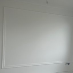 BUD-MAR USŁUGI BUDOWLANE MARCIN MAŃKOWSKI - Biała ściana z dekoracyjną ramą i listwami przypodłogowymi, widoczne gniazdka elektryczne, surowy stan wykończenia.